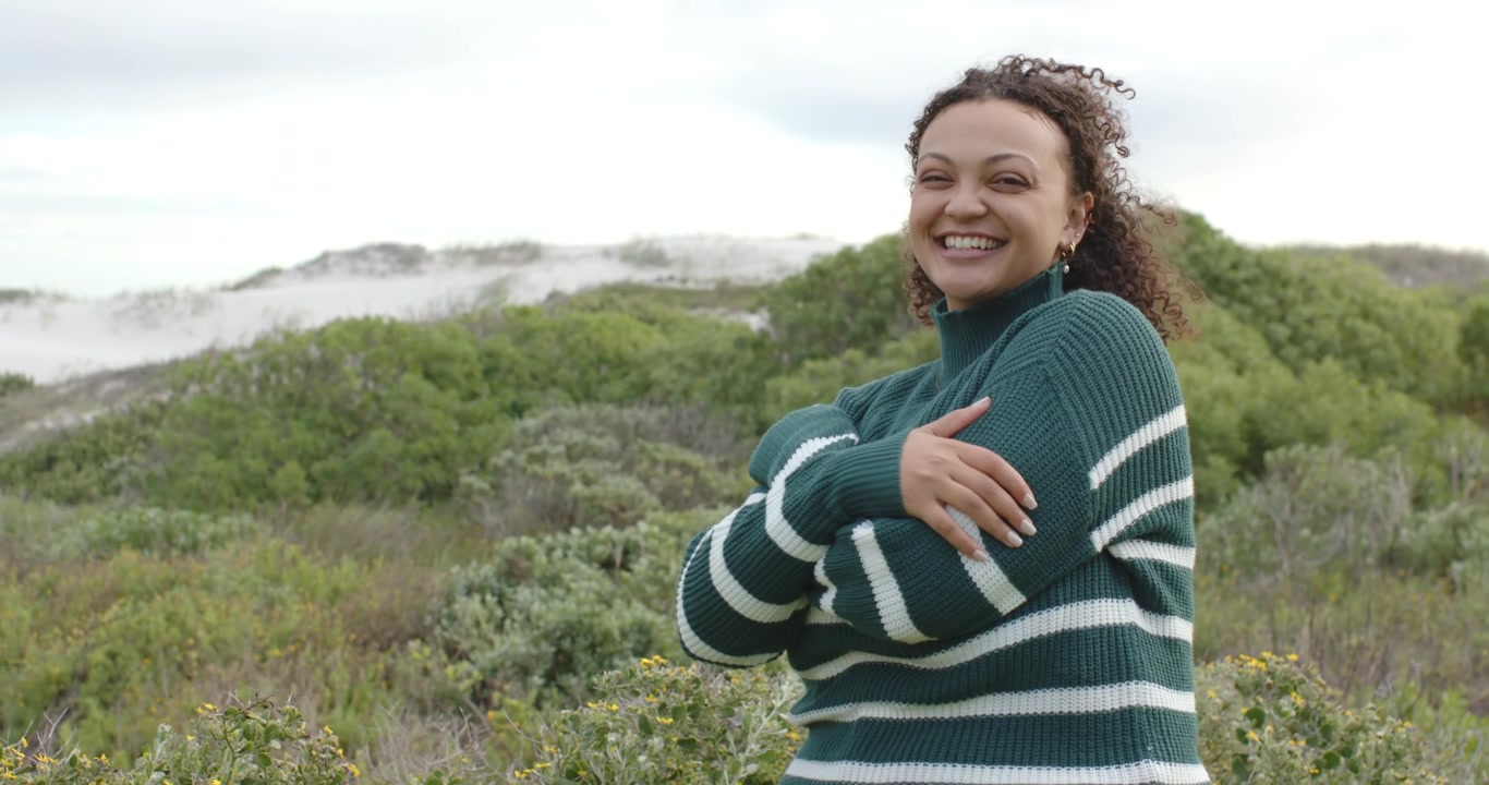 Donna con le braccia incrociate di fronte alla telecamera sulle dune, con maglia verde, il vento che le solleva i ricci mentre cerca calore, con spazio per il testo.