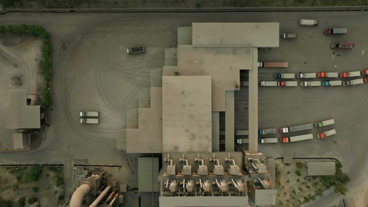 Top-down real time view of a cement factory with parallel truck lanes and industrial structures framed by paved lots and dust trails, aerial view, drone shot