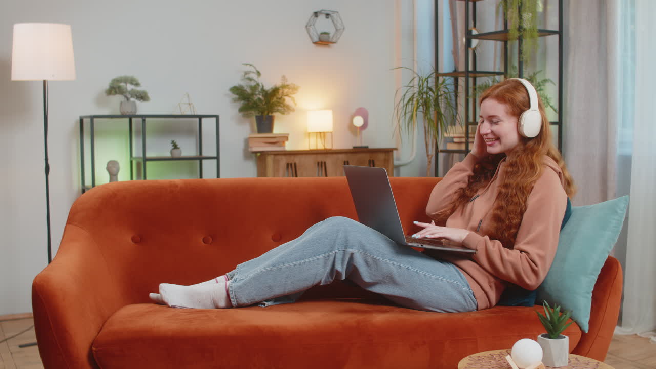 feliz y relajada joven caucásica con auriculares inalámbricos escuchando música usando una computadora portátil en el sofá