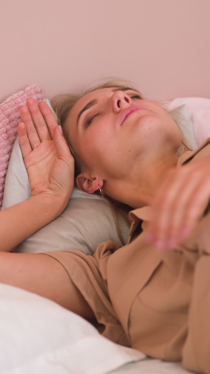 Sleepy dressed blonde woman wakes up on bed and covers head with soft white pillow avoiding bright daylight and noise at home slow motion