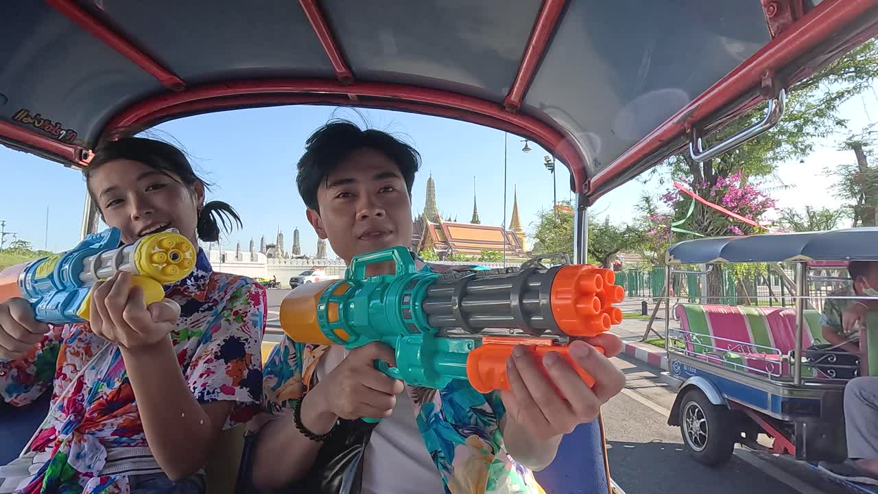 คู่รักที่มีความสุขสวมเสื้อลายดอกไม้เล็งปืนฉีดน้ำจากรถตุ๊กตุ๊กที่กำลังเคลื่อนที่ในกรุงเทพฯ