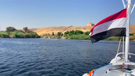 egyptian flag waving on the bow of a boat sailing down the nile with a view of a temple in the desert