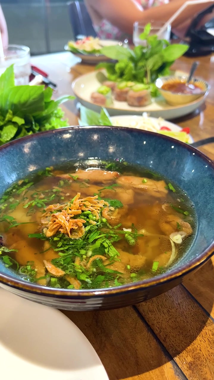 Một tô phở gà hấp dẫn được trang trí bằng rau thơm, chụp trong không khí nhà hàng sống động với ánh sáng tự nhiên.