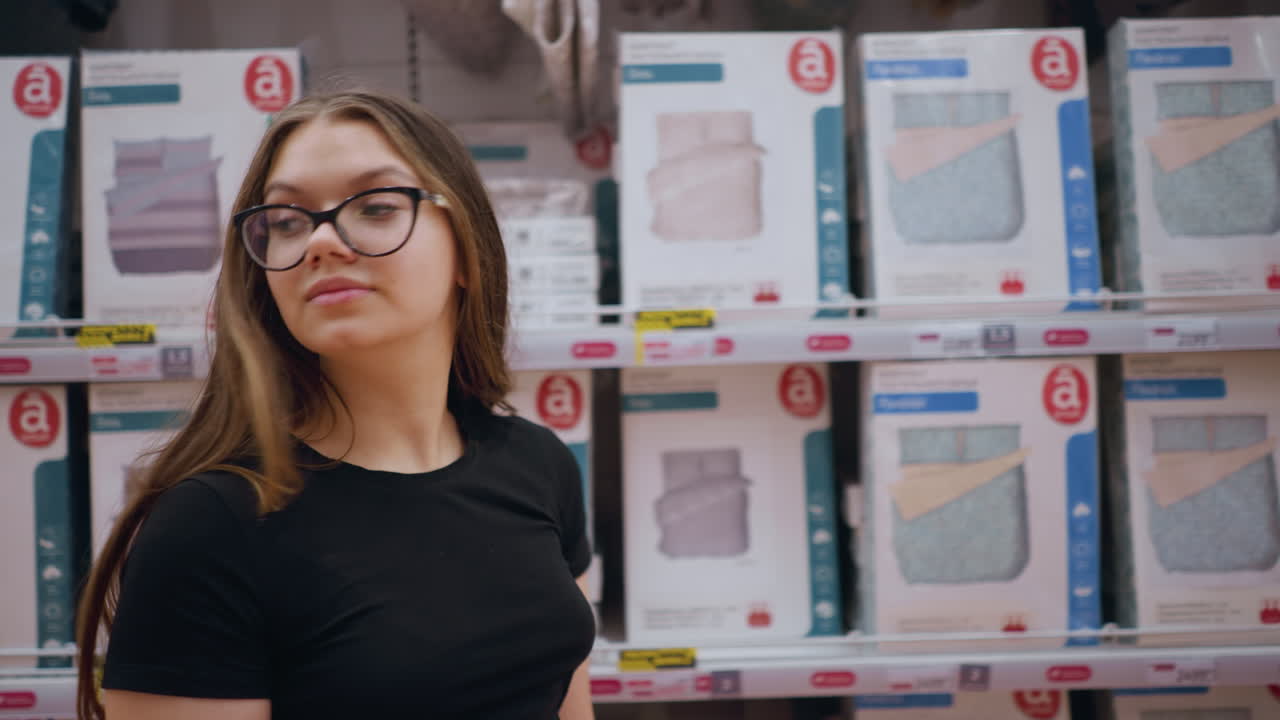 mujer joven caminando más allá de la estantería de la tienda abastecida con productos de cama, ella mira a su alrededor, examinando diferentes opciones de edredones y almohadas, el entorno de la tienda con textiles para el hogar cuidadosamente dispuestos