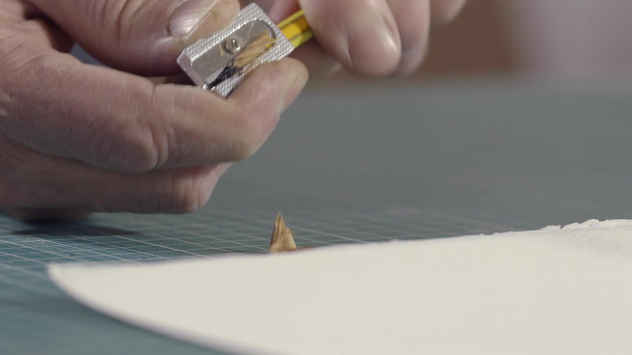 Primer plano de manos afilando un lápiz de madera con un sacapuntas manual. Las virutas de lápiz caen sobre la superficie de trabajo, simbolizando preparación, creatividad y artesanía.