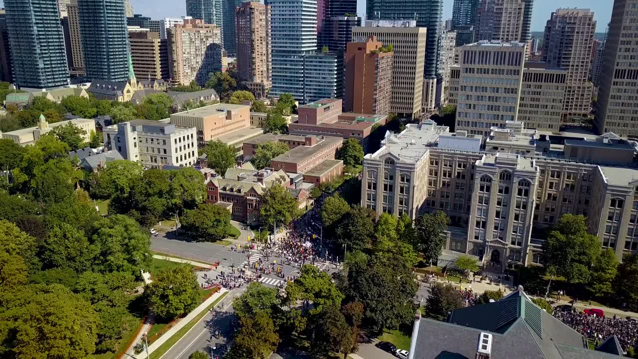 Epic Aerial Drone Pull Back Reveal of Protest March, Downtown Toronto. Crowd of people protesting march on city streets among huge office buildings and condo towers, wide daytime exterior in 4k