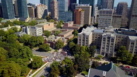 epic aerial drone pull back reveal of protest march, downtown toronto. crowd of people protesting march on city streets among huge office buildings and condo towers, wide daytime exterior in 4k