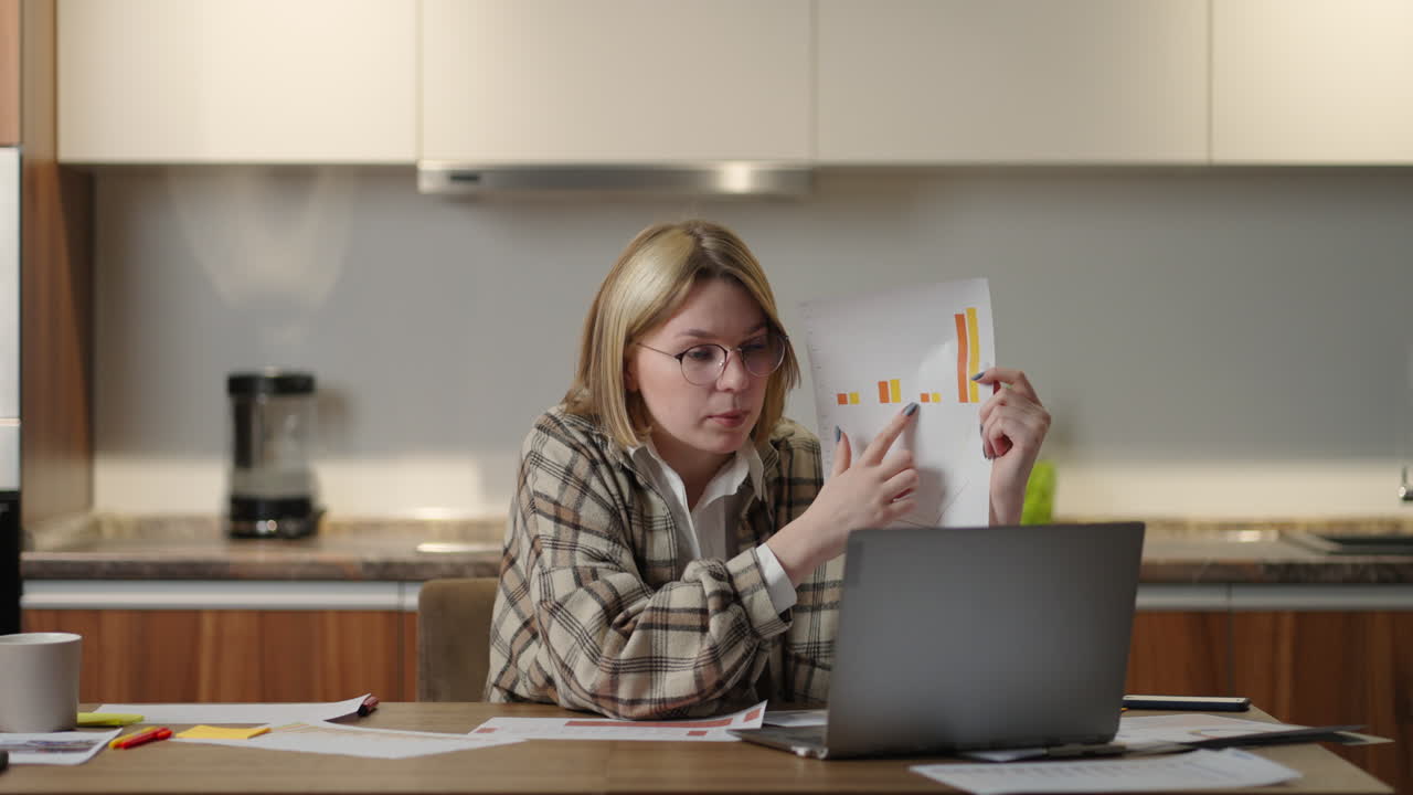 una mujer joven con gafas en casa muestra un gráfico a una cámara portátil mientras está sentada en casa en la cocina. trabajo remoto de la oficina en el hogar. llamada de video y conferencia con gráficos y tablas.