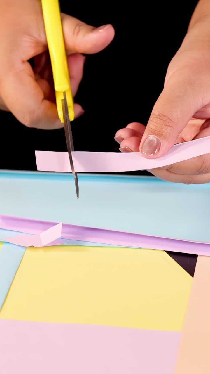 Hands use yellow scissors to cut pastel paper strips on black background, bright studio lighting