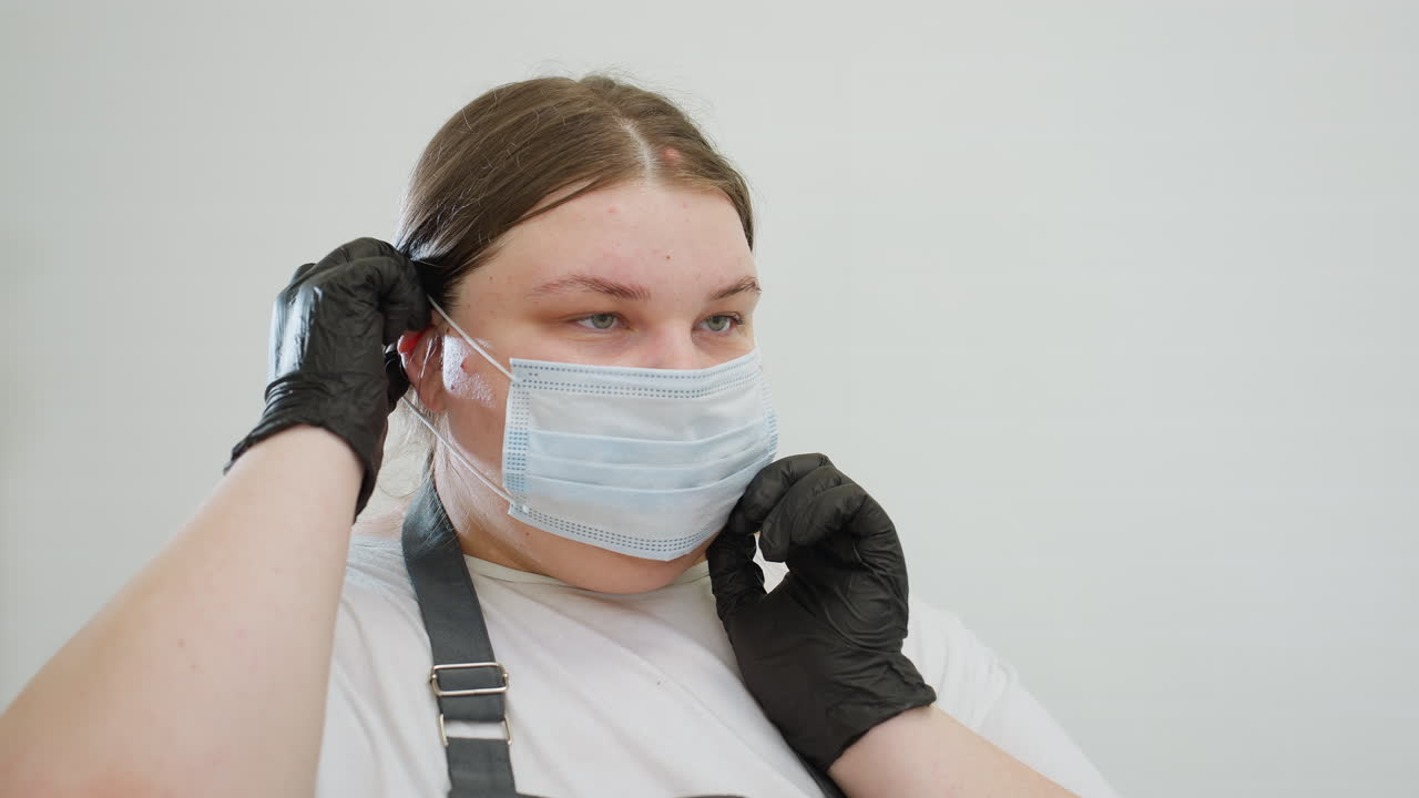 Beautician with blond hair carefully adjusts protective face mask while wearing black gloves in clean white workspace. Focused on hygiene and preparation before attending to client session