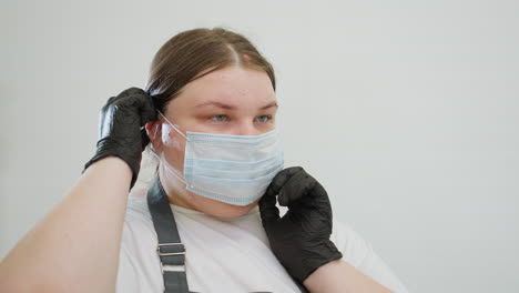 beautician with blond hair carefully adjusts protective face mask while wearing black gloves in clean white workspace. focused on hygiene and preparation before attending to client session