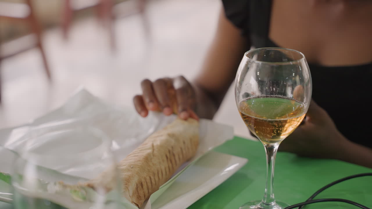 Mulher desembrulha um doce ao lado de uma taça de vinho, mãos levantam um recipiente plástico enquanto a bebida dourada brilha na taça, close-up espontâneo de um momento de refeição, vestido casual e postura relaxada, mesa de café urbano.