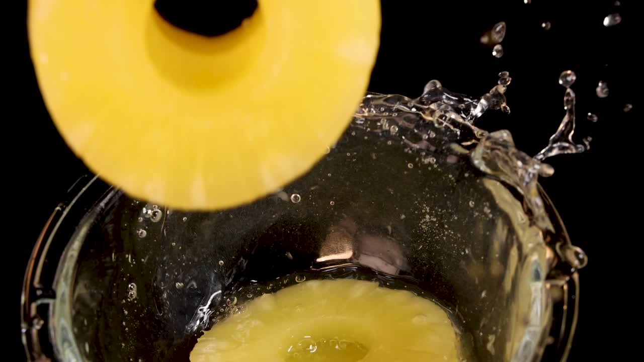 Sliced pineapple rings fall into a clear glass bowl, creating a splash against a black background with dramatic studio lighting and a top-down camera angle