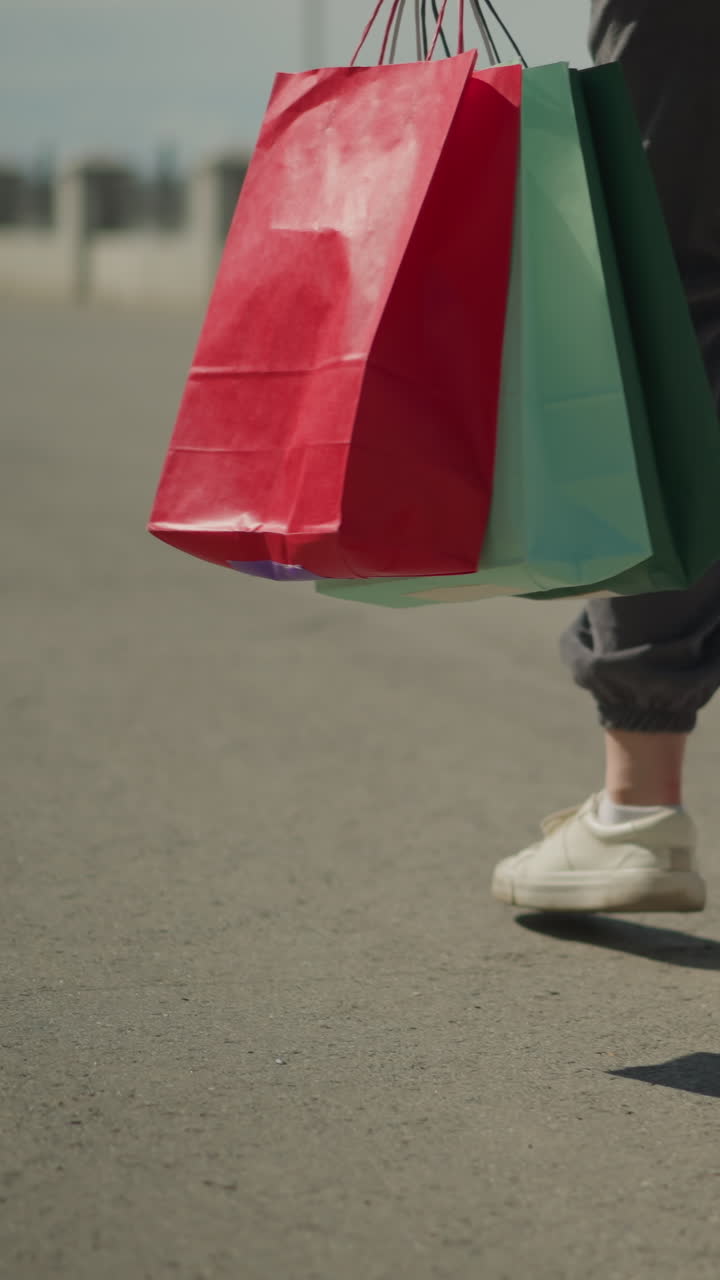 빨간색, 초록색 및 민트색 쇼핑 가방을 손에 들고 도로를 고 있는 사람의 다리 시각, 멀리서 자전거를 타고 있는 사람을 특징으로 하는 흐릿한 배경