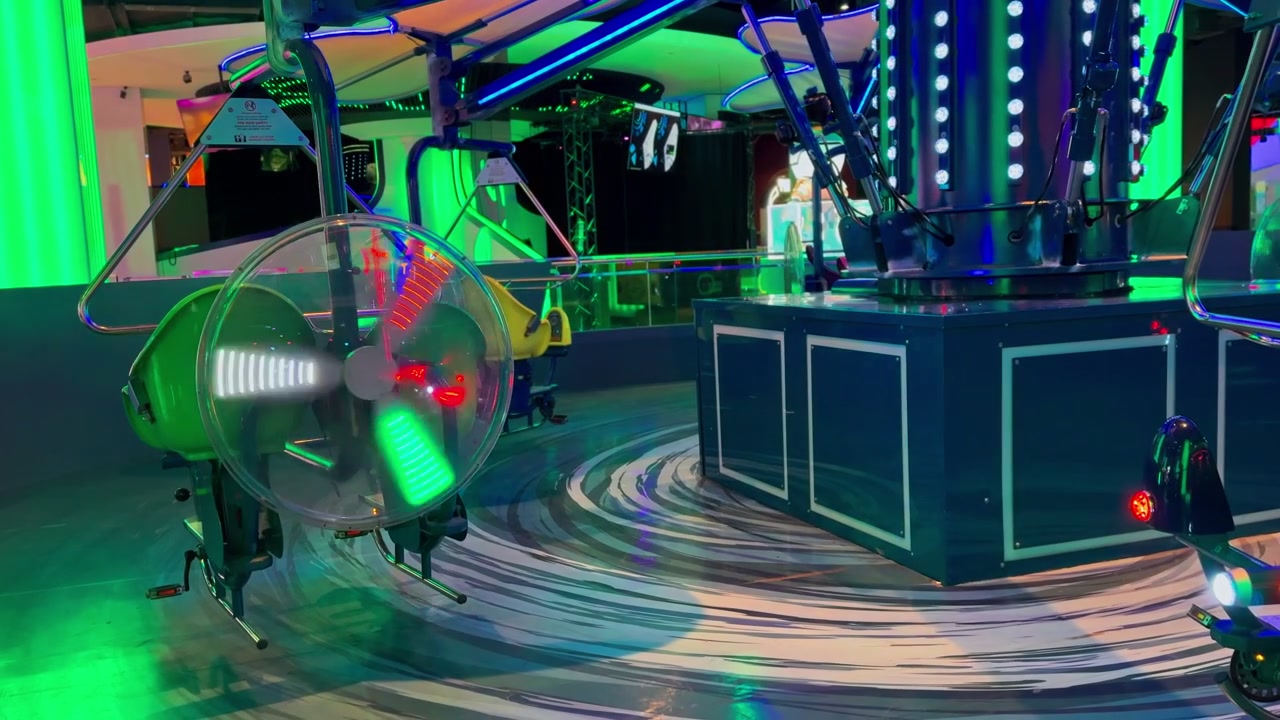 Un'inquadratura ampia e statica di un'attrazione girevole vuota e colorata all'interno di un parco divertimenti al coperto e buio. L'attrazione meccanica presenta luci LED al neon verdi e blu luminose e un'elica posteriore rotante.