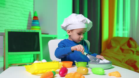 beautiful baby boy in blue sweater and white chef cap sits at table. cute little cook uses spatula and toy frying pan with eggs.
