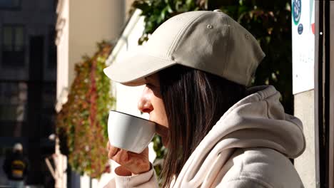 girl in white hoodie and cap peacefully drink cup facing sunlight in cafe