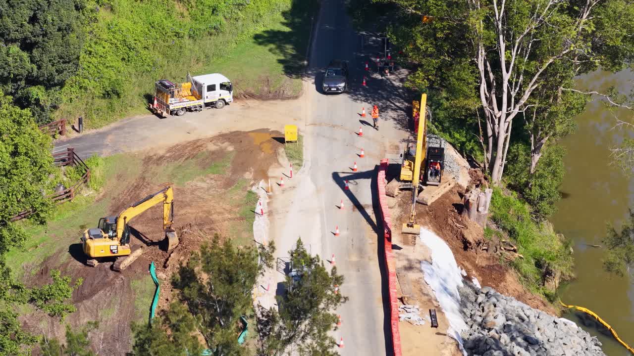 Drone footage captures roadwork with excavators and vehicles near a river in Uki, NSW, Australia under bright daylight