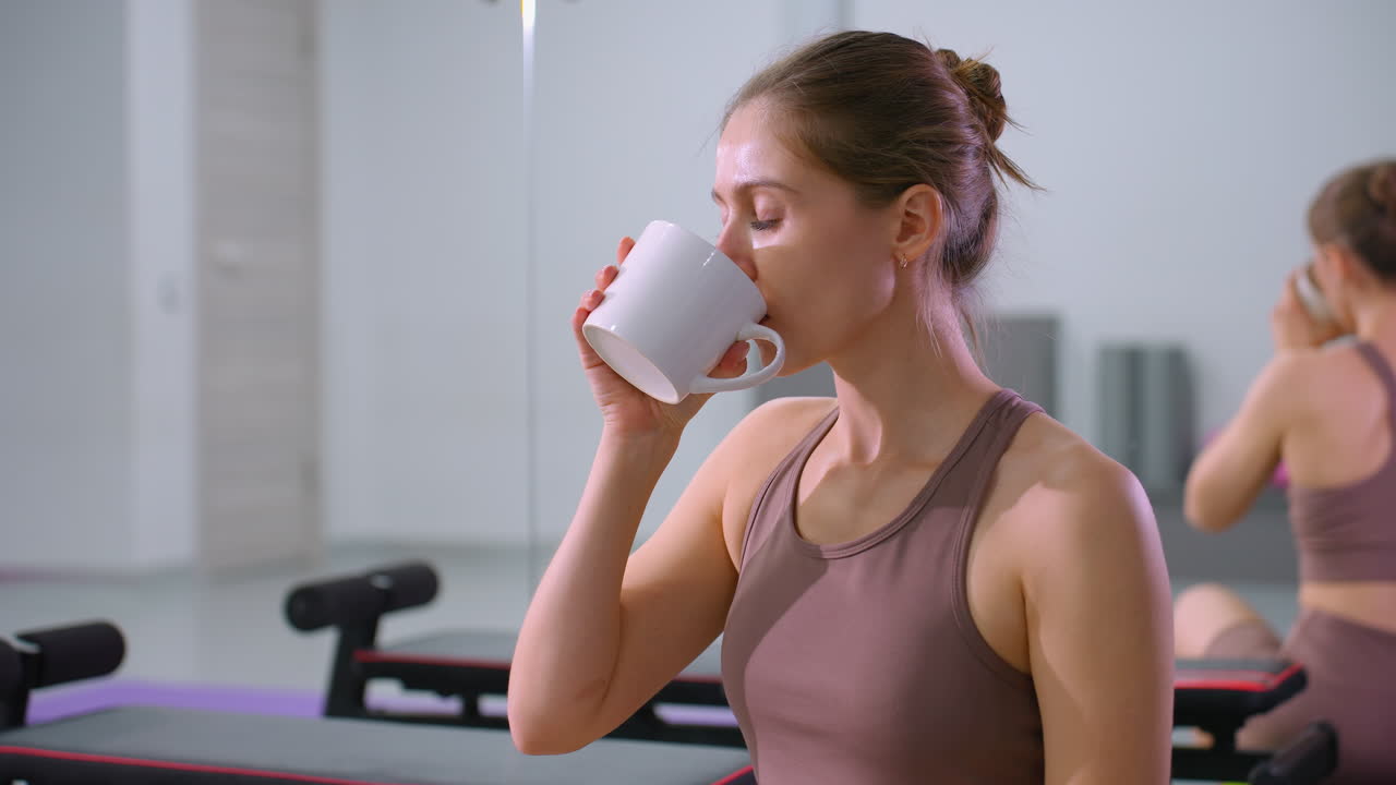mujer toma un descanso de la sesión de fitness, bebiendo café mientras está sentada en el gimnasio, un gran espejo refleja una postura relajada, equipos de entrenamiento y esteras en el fondo