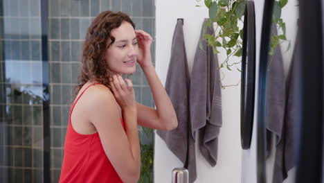 applying skincare, woman in red top looking in bathroom mirror, smiling