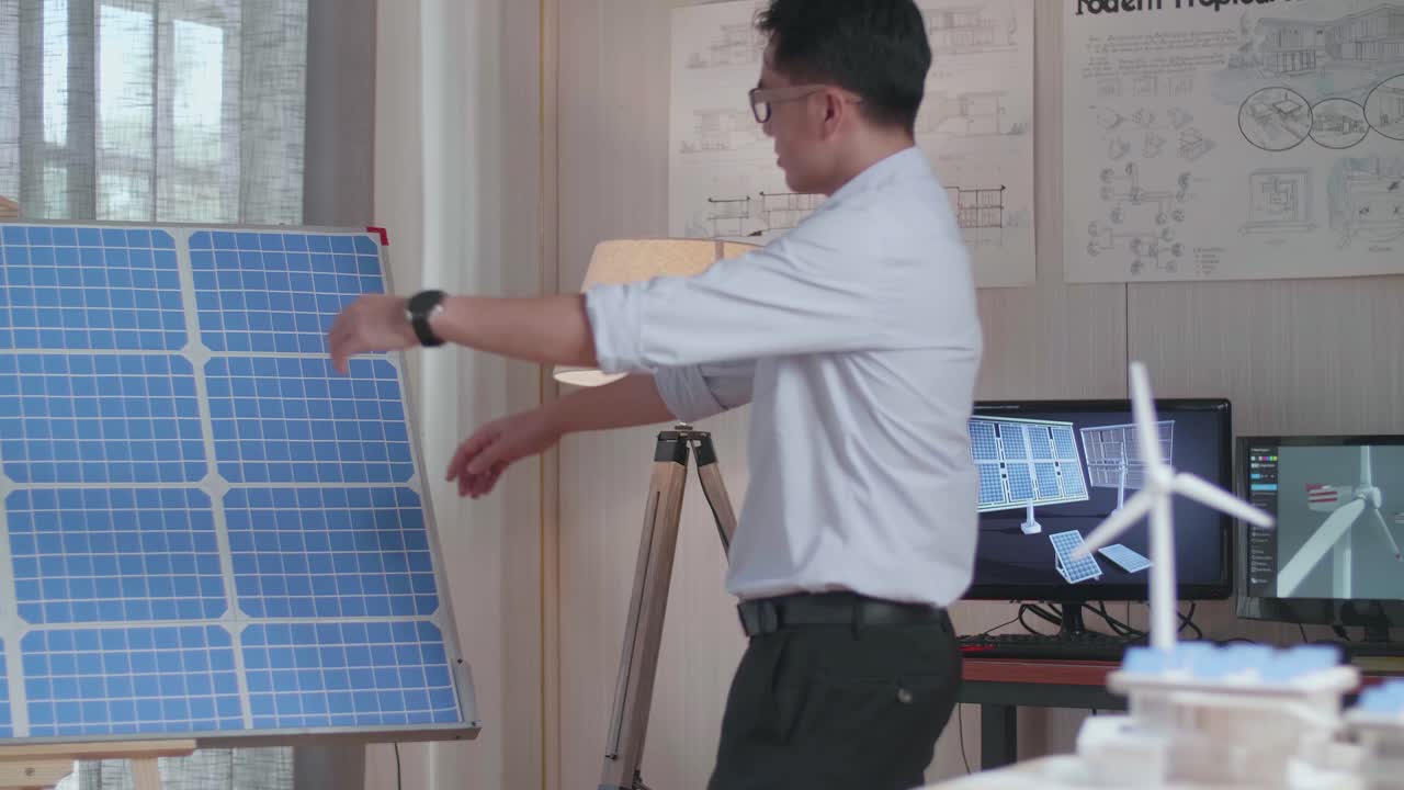 아시안 남자가 태양 전지를 보며 걸어 들어가는데, 그 방에는 모형 태양 전지판, 작은 집, 그리고 테이블 위에 풍력 발전기가 있다.