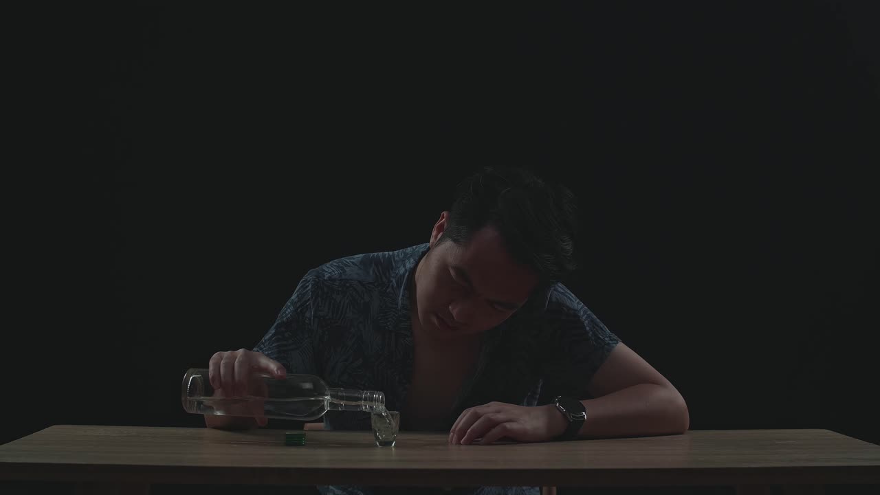 술에 취한 우울한 아시아 남자가 술을 마시기 전에 글라스에 보드카를 부어 주고 검은색 바탕에 테이블을 르는 주먹을 사용합니다.