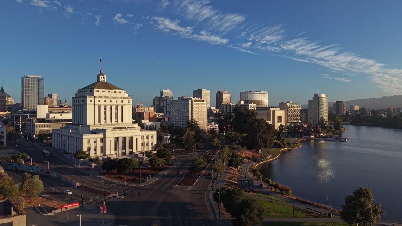 Luce dorata del mattino che illumina il tribunale della contea di Alameda e lo skyline del centro di Oakland accanto alle acque del Lago Merritt, con nuvole sottili che si muovono attraverso un cielo azzurro limpido. Ripresa con drone in ascesa.
