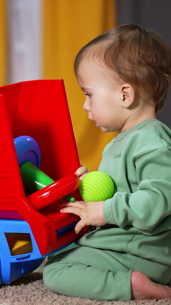 Sweet child playing with a bright lorry on the soft carpet. Toddler turns the car to take the toys out of the car basket. Blurred backdrop. Vertical video