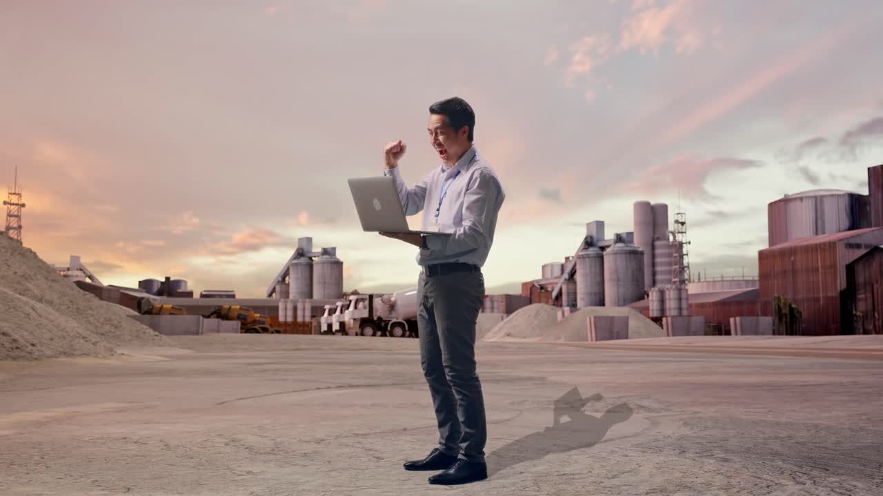 Vista lateral de cuerpo entero de un trabajador profesional asiático de pie con su ordenador portátil en una planta de cemento industrial al atardecer, levanta el puño con un grito de victoria después de escribir en el portátil.