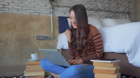 estudiante asiática graciosa sentada en un piso con su computadora portátil rodeada de libros, feliz de que finalmente resolvió la tarea en su computadora