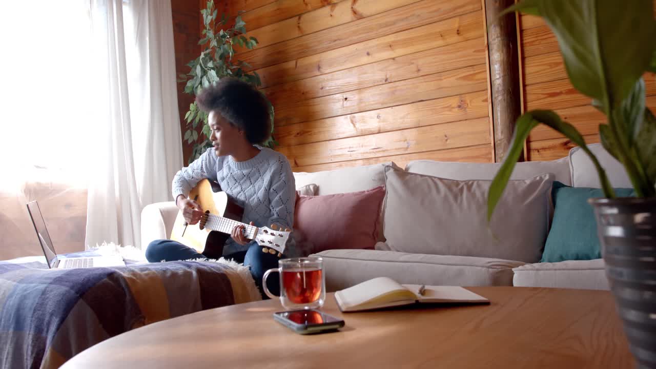 Happy african american woman playing guitar using laptop in living room, in slow motion