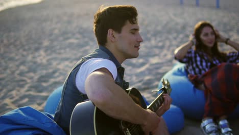 Junger-Mann-Spielt-Gitarre-In-Einer-Gruppe-Von-Freunden,-Sitzt-Auf-Sesseln-Am-Strand-Und-Singt-An-Einem-Sommerabend-Bei-Sonnenuntergang.-Zeitlupenaufnahme.