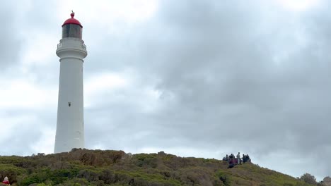 a lighthouse stands tall against a cloudy sky, surrounded by greenery and distant visitors, captured in a serene coastal setting