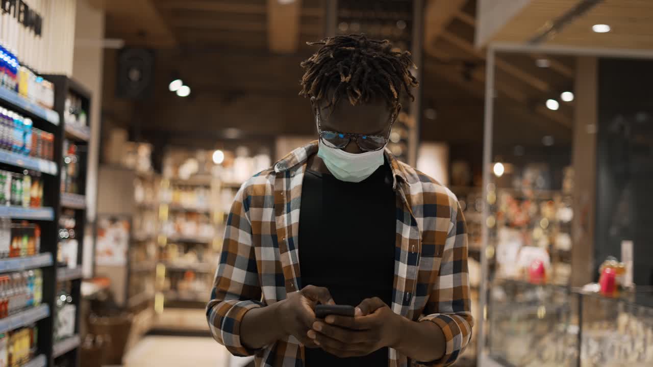 African American man in mask busy with smartphone walks at the store