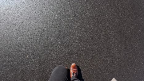 first-person view of a person walking on dark smooth asphalt in bright sunlight, wearing brown leather shoes and dark pants.