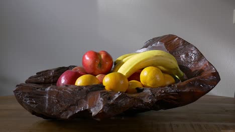 a male hand takes a banana, and then an apple from a wooden bowl of fruit standing on a wooden table