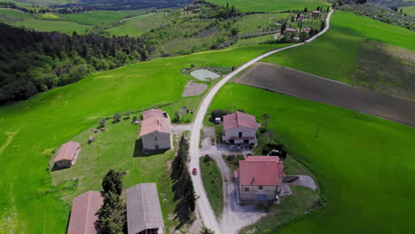 Car-on-road-by-houses-and-green-fields-in-Tuscany,-wide-aerial-reveal