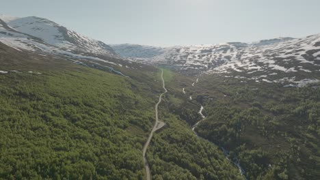 a winding mountain road in the lush green valley with snow-capped peaks in norway, aerial view