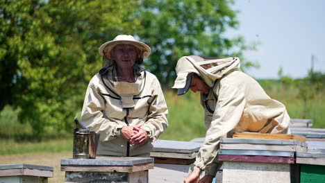 smiling adult men working at bee farm. younger apiculturist putting frame into a hive. multiple bees fly around. blurred nature backdrop.