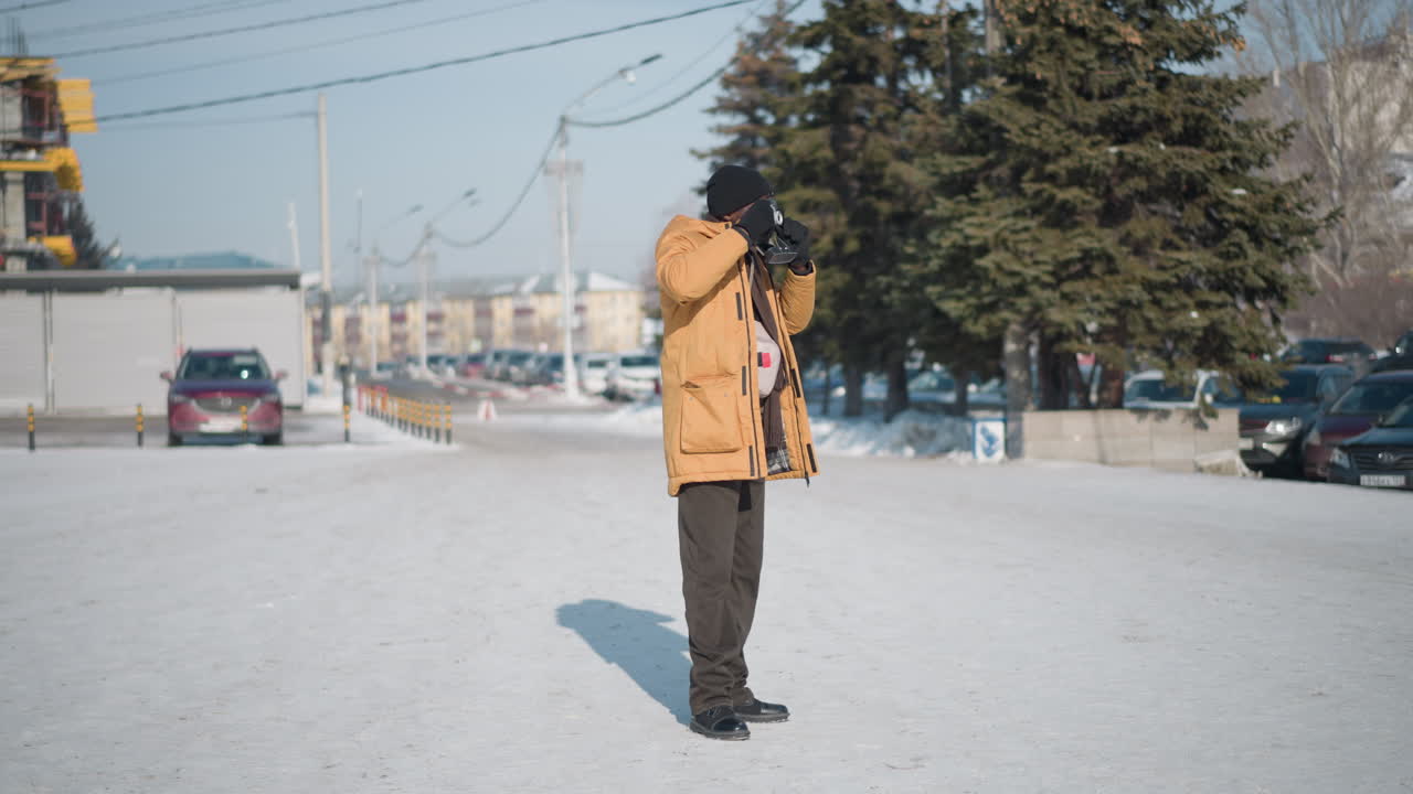 back view outdoor photographer in yellow coat capturing cars parked on snowy city plaza amid trees and buildings blur background using camera on strap with lens under soft winter daylight backdrop