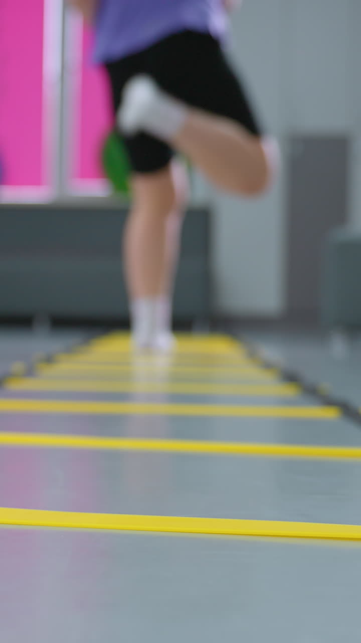vista trasera de la dama de púrpura saltando escalera de agilidad para la velocidad y la coordinación mientras otro niño en el fondo borroso trabaja en el equilibrio, varios equipos de gimnasio y herramientas de entrenamiento visibles