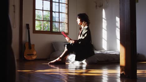 Relaxed-mixed-race-woman-sitting-on-bed-reading-book-in-sunny-cottage-bedroom
