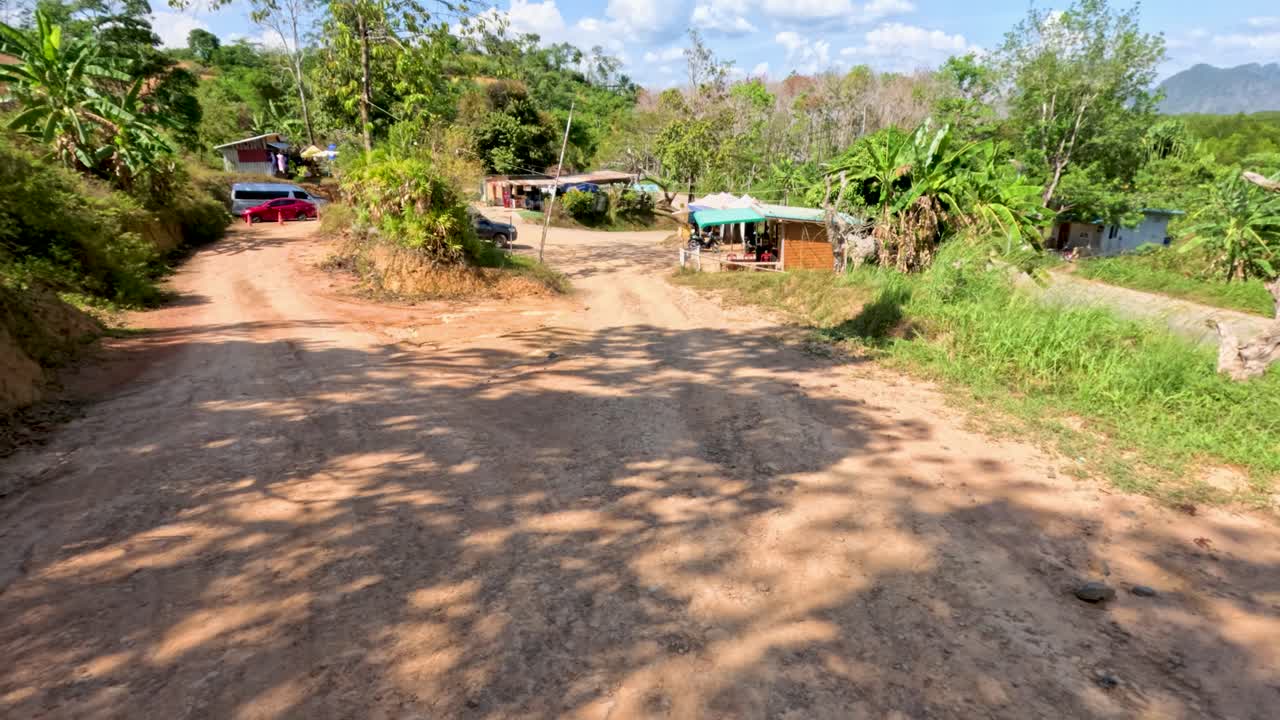 Vehicle travels along sunlit dirt road past roadside stalls, lush greenery, and parked cars, daytime