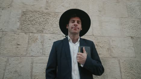 un hombre con trajes tradicionales de pie con confianza contra una pared de piedra, sosteniendo un libro