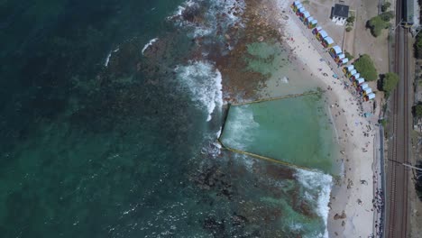 Top-down-shot-from-drone-of-train-tracks-next-to-ocean-with-tide-pool,-coloured-beach-shacks-on-beach