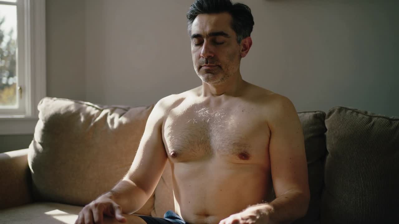 Uomo adulto seduto che respira sul divano vicino alla finestra, con la luce del sole che si sposta sul petto nudo e sui pantaloncini.