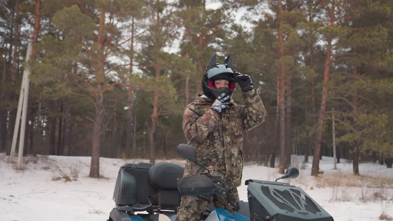 Pilota mimetizzato accanto all'ATV prepara l'attrezzatura in una foresta di pini innevata, regolando casco e occhiali mentre indossa giacca mimetica invernale e guanti, in silenziosa attesa prima di una corsa fuoristrada estrema.