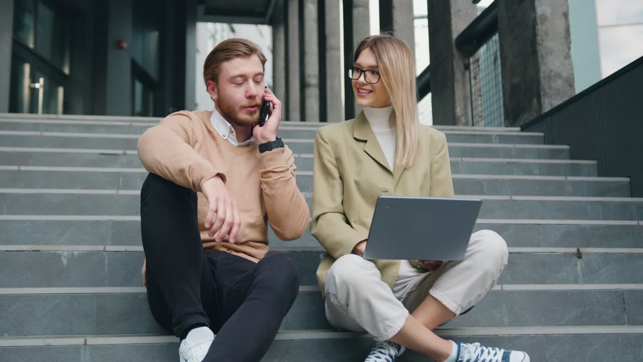 Attractive man and woman sitting on stairs in urban city center working on laptop, talking on mobile phone smiling, stylish freelance people using technology outside, making notes. Outside