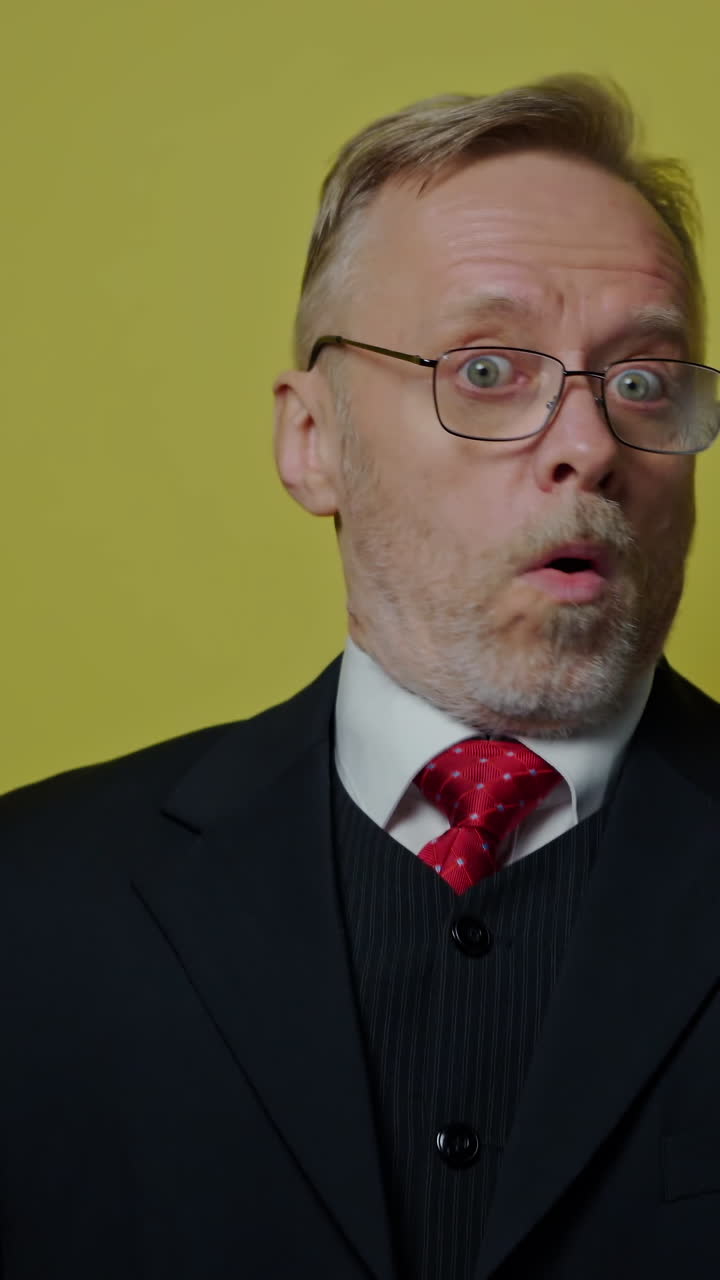 Positive businessman showing different emotions. Mature man in glasses and elegant costume shows funny face on the camera on yellow studio background. Vertical video