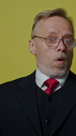 positive businessman showing different emotions. mature man in glasses and elegant costume shows funny face on the camera on yellow studio background. vertical video